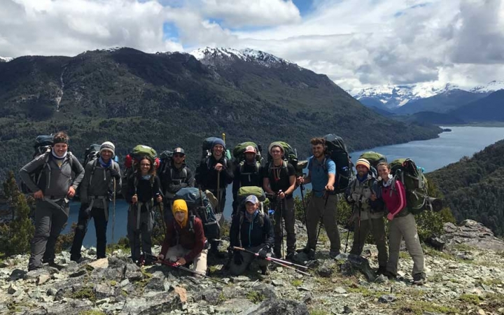 A group of people wearing backpacks pose for a group photo on an overlook. Behind them there are mountains and river. 